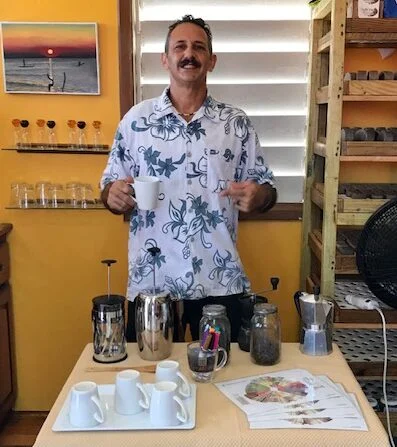 A man in a floral shirt holds a mug behind a table of coffee items and cups, set against a yellow wall with a sunset picture, enjoying the Beach-side Coffee Experience.