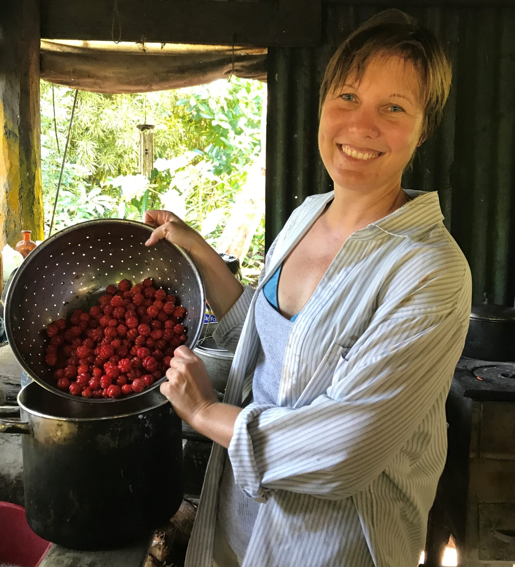 A smiling person holds a colander full of red berries over a large pot in a rustic kitchen setting.