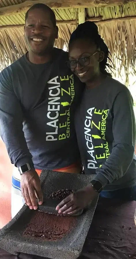 Two people in "3 Adventures Combo Tour" shirts stand together, working with cocoa beans on a flat surface in a rustic setting.