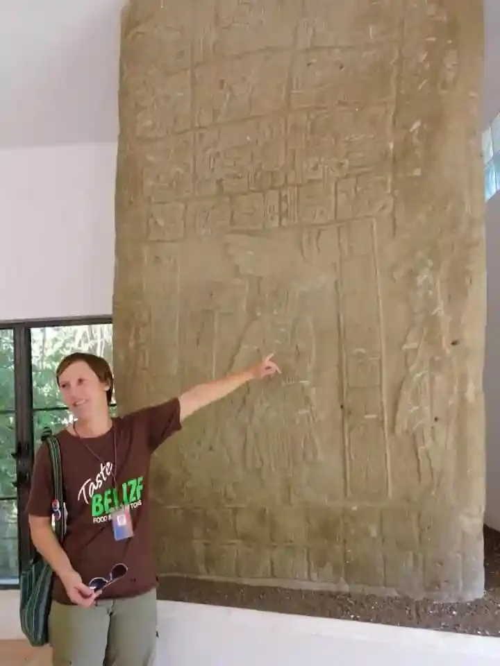 A person points at an ancient stone slab with carvings, displayed indoors. They wear a "Chocolate & Nim Li Punit Maya Site" shirt and hold sunglasses.